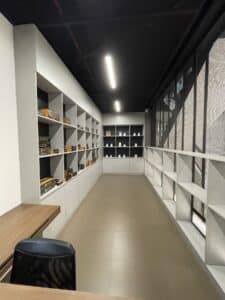 A modern, well-lit room with shelves displaying various items, including books and boxes. A wooden table and a black chair are positioned in the foreground. The space features large windows on the right side, perfect for either residential comfort or commercial use after a stylish renovation.