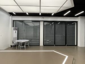 A modern, empty commercial space with a hexagonal ceiling light, tiled floor, a round table with four metal chairs, and a wall of large glass doors with closed blinds. A standalone air conditioner is near the wall, reflecting a thoughtful design and build approach for an office setting.