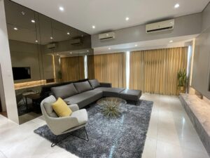 Modern living room with a grey L-shaped sofa, a light grey armchair with a yellow cushion, glass coffee table, large grey rug, mirrored wall, and beige curtains. The space, ideal for both residential and office settings after renovation, features ceiling lights and air conditioners.