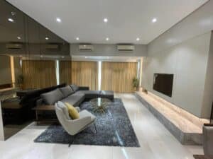 Modern living room with a gray sectional sofa, a single armchair with a yellow pillow, and a glass coffee table. The space features an interior fit-out including a large area rug, wall-mounted TV, and floor-to-ceiling windows with beige curtains.