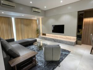 Modern living room with a large sectional sofa, armchair, and glass coffee table on a shaggy rug. Wall-mounted TV on a sleek entertainment unit. Large windows with beige curtains and recessed lighting showcase the quality of interior fit-out typical in both residential spaces.