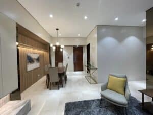 Modern residential dining and living area with a wooden dining table and chairs, a gray upholstered armchair with a yellow pillow, white tile flooring, and contemporary light fixtures.