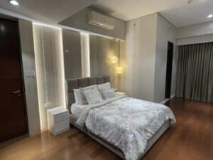 A modern bedroom with a double bed, white linens, bedside table, hanging pendant lights, and an air conditioner above the headboard. The tastefully renovated space features light-colored walls and wooden flooring, perfect for residential comfort.