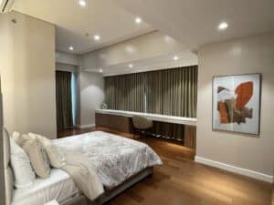 A modern bedroom with wooden flooring, a large bed with patterned bedding, a study desk with a chair, and a painting on the wall. This beautifully renovated space is illuminated by recessed lighting, showcasing expert interior fit-out craftsmanship.