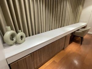 A long white countertop with two cylindrical vases and a chair stands against a backdrop of tall, pleated curtains on a wooden floor, showcasing an elegant office interior fit-out.