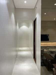 A modern indoor hallway with white walls and a tiled floor, part of an impeccable interior fit-out, leads to a lit living room area in the background. The living room contains a TV mounted on the wall, a coffee table, and a sofa.