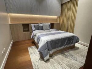 A neatly made bed with gray and white bedding is in a modern bedroom with beige curtains, wooden flooring, and a soft area rug. The headboard features built-in shelves and warm lighting. The room reflects the precision of an interior fit-out typically seen in high-end commercial spaces.