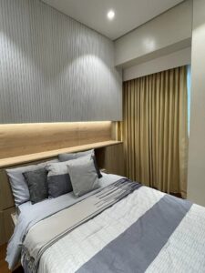 A modern bedroom with a neatly made bed, beige accent wall, built-in wooden headboard with lighting, and beige curtains covering a window showcases a superb residential renovation. The thoughtful interior fit-out enhances the cozy yet stylish ambiance of the space.