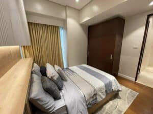 A modern bedroom with a neatly made bed, beige curtains, wooden closet, and recessed lighting. The walls and ceiling are white, and a light-colored area rug is partially visible on the wooden floor. The room's interior fit-out exhibits a sleek professionalism reminiscent of a commercial office space.