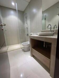 A modern office bathroom with a glass-enclosed shower, toilet, and vanity sink. The walls and floor are tiled in neutral tones for a sleek commercial look, with a mirror above the vanity.