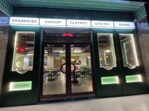 A restaurant entrance with a green exterior, glass doors, and a neon "welcome" sign boasts a fresh design and build. Overhead signs display menu items: Spareribs, Dim Sum, Claypot, Stir Fry, and Siomai. Tables and chairs are visible inside, evoking an atmosphere more akin to a lively office than traditional dining.