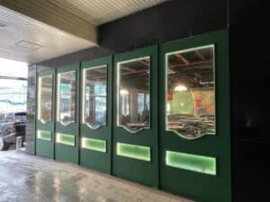 A green storefront with large glass windows and illuminated panels, viewed from the street. Cars are parked nearby, and the interior fit-out reveals a dining area with visible seating arrangements.