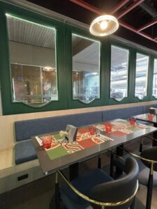 A restaurant table set with placemats, red glasses, and napkin holders on a grey tabletop, complemented by blue cushioned seating and large green-framed windows in the background reflects a meticulously designed interior fit-out.