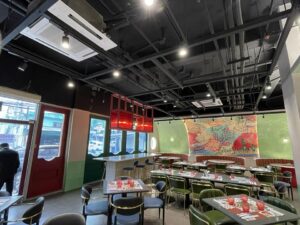 A modern restaurant interior with tables set, green chairs, and wall art. Exposed ceiling with lights and visible ventilation add a chic touch to the commercial space. Large windows brighten the room, while red accents on walls and fixtures create a vibrant atmosphere.