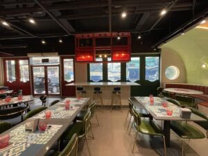 A cozy restaurant interior with neatly arranged tables and chairs, colorful place settings, and a bar area with stools. Following a thoughtful renovation, circular windows and a mix of red, green, and beige colors bring warmth to this charming commercial space.