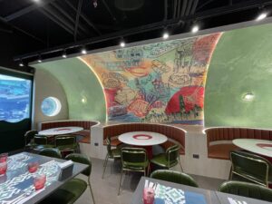 A modern restaurant interior, designed and built with care, features round tables with green chairs, red glasses, and a vibrant mural depicting various scenes and dishes on a curved wall. The refined atmosphere hints at a blend between chic residential spaces and dynamic office environments.