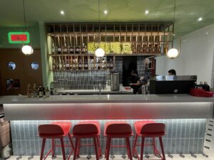 A modern bar with red stools, a variety of bottles on shelves behind, and two staff members working behind the counter displays a flawless design and build. Neon signs and ambient lighting add to the cozy atmosphere.