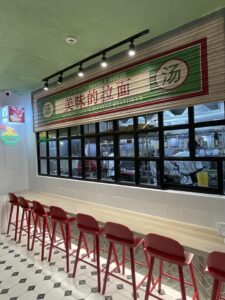 A newly renovated noodle shop with a counter lined with red stools faces an open kitchen. A sign above the counter reads "Ramen Noodle House" and "Delicious Ramen" in Chinese. Geometric floor tiles and green walls add to the inviting atmosphere of this vibrant commercial space.