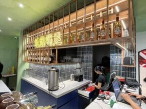 A bartender works behind a counter at a bar with a lit shelf displaying various bottles. A neon sign in a non-English language is seen above the shelf, showcasing the stunning design and build of this newly renovated space.