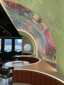 A modern cafe interior with curved booths, a wooden table, and a large mural on the curved ceiling. Natural light enters through a round window and glass doors in the background, giving it the feel of an office that's undergone a stylish renovation.