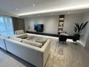 A modern living room features a large white sectional sofa, a wall-mounted TV, a lounge chair, and minimalist decor. Designed and built during an extensive renovation, the space is illuminated by recessed lighting and natural light from large windows.