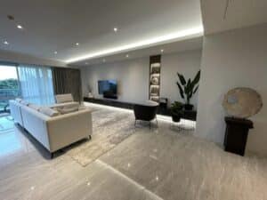 A modern living room with a white sofa, a black armchair, plants, a wall-mounted TV, and a decorative stone disc. The floor is made of marble tiles, and large windows offer a view outside. This interior fit-out exemplifies elegant design and build quality throughout the space.