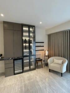 A modern living room with a tall cabinet featuring glass doors, a gray armchair with a pillow, a small side table with a lamp, and beige floor tiles. A window with closed beige curtains is to the right, showcasing impeccable interior fit-out akin to an upscale office's design and build.