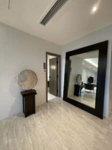 A modern interior hallway with beige marble flooring, a large black-framed mirror, a decorative round stone piece on a pedestal, and an open door leading to another room showcases meticulous design and build renovation.