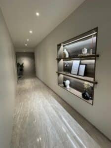 A modern hallway with marble floors, recessed lighting, and a built-in shelf displaying decorative items including framed pictures, vases, and a plant showcases an exquisite interior fit-out suited for both residential and commercial spaces.