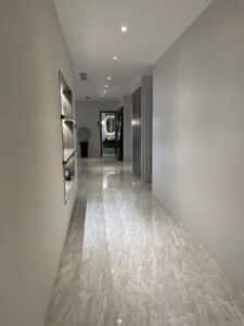 A modern hallway with marble flooring, recessed lighting, and built-in shelving on the left. An office door at the end opens to a room with a large mirror and decor, showcasing a seamless interior fit-out.