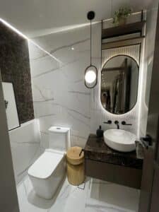 A modern bathroom with marble walls, a toilet, a round mirror above a dark vanity with a white sink, a cylindrical light fixture, and a wooden stool showcases an impressive interior fit-out.