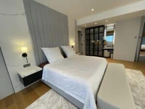 A modern bedroom with a large bed, white bedding, and an upholstered headboard. The residential space features bedside tables with wall-mounted lamps, a bench at the foot of the bed, and a sliding glass door leading to another room.