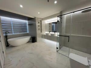 A modern bathroom with a freestanding bathtub, double sink vanity, glass-enclosed shower, toilet, and marble flooring. The wall has horizontal striped patterns. The room is well-lit with overhead lights, reminiscent of a sleek commercial interior fit-out that could easily grace an upscale office.