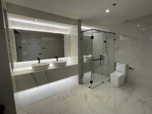 A modern bathroom with marble tiles, featuring a double sink vanity with illuminated mirrors, a glass-enclosed shower, and a toilet showcases the meticulous interior fit-out design often found in high-end commercial spaces.