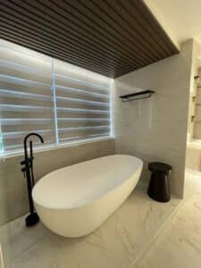 A modern residential bathroom features a freestanding white bathtub, black floor-mounted faucet, and small black stool. The space boasts marble walls and floors, with a window adorned with horizontal blinds—a perfect blend of thoughtful design and build.