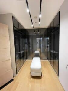 A modern residential walk-in closet with black glass-fronted cabinets, a white upholstered bench in the center, and wooden flooring.