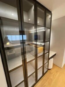 A modern bedroom features a glass wardrobe with multiple compartments and a built-in desk beneath it. The interior fit-out includes a large window and sleek wooden flooring, exemplifying stylish design and build principles.