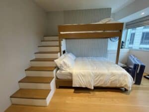 A residential bedroom with a wooden bunk bed; the lower bed is a double and the upper bed is a single. Steps to the upper bunk are on the left. A window is on the right side, allowing for natural light to flood the room, showcasing an elegant interior fit-out.