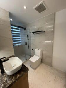 Modern residential bathroom with marble tiles featuring a walk-in shower, toilet, vessel sink on a dark countertop, and a window with horizontal blinds.