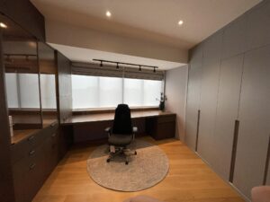 A modern home office, recently renovated, boasts a large window, built-in cabinets, a desk with a black office chair, and a circular rug on a wooden floor.