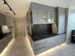 A modern kitchen, perfect for a home office renovation, features grey cabinets, marble countertops and backsplash, under-cabinet lighting, and tile flooring. A door is visible in the background.