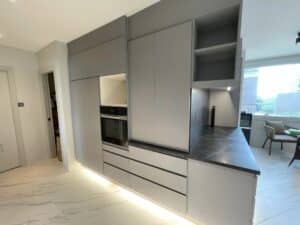 Modern kitchen with minimalist grey cabinetry, built-in oven, under-cabinet lighting, and dark countertops. A recent renovation highlights the sleek design and build quality. The window in the background overlooks a cityscape.