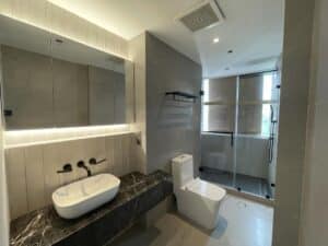 Modern bathroom with a black marble countertop, white sink, wall-mounted faucet, toilet, and glass shower enclosure. Recessed lighting above the mirror and a frosted window in the shower area make it ideal for both residential and commercial renovations.