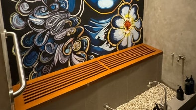 A bathroom with a wall-mounted wooden shelf, pebble flooring under the sink area, and a colorful floral mural on the wall, exemplifies exquisite interior fit-out suitable for both commercial and residential spaces.