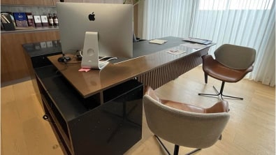 A modern office, the result of a recent renovation, features a sleek wooden desk, an iMac computer, and a wooden chair on the left. A gray chair sits on the right, positioned in front of a window with white curtains. Books and a plant adorn the shelf, showcasing thoughtful design and build elements.