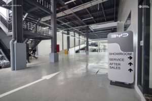 Modern indoor car dealership area with a BYD sign pointing to showroom, service, and after sales sections; stairs and open space visible.