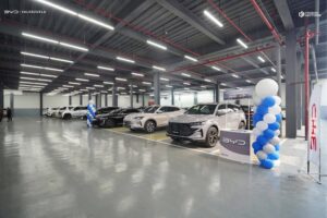A row of new cars is parked indoors in a well-lit showroom with high ceilings. Blue and white balloons and banners are displayed nearby.