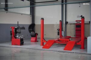 A car service bay with red vehicle lift, tire changer, wheel balancer, and tool cabinet in a clean, modern garage.