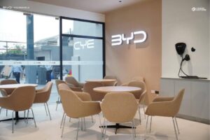 Modern showroom lounge with beige chairs and round tables, BYD logo illuminated on the wall, and a charging device mounted nearby. Large glass windows provide outdoor visibility.