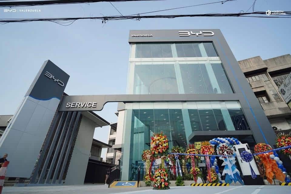 A modern BYD dealership and service center with glass facade, BYD logos, and flower arrangements at the entrance on a clear day.